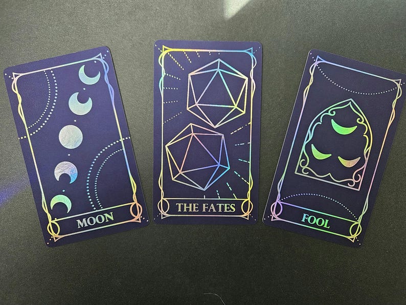 May include: Three rectangular cards with a dark blue background and holographic foil details. The first card shows moon phases and the text MOON. The second card features two icosahedron dice and the text THE FATES. The third card displays a gothic arch with crescent moons and the text FOOL.