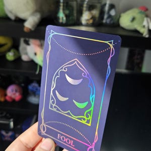 May include: A rectangular tarot card with a dark purple matte finish and holographic foil detailing. The card features an arched window design containing three crescent moon shapes, with the word FOOL printed in a rectangular frame at the bottom.