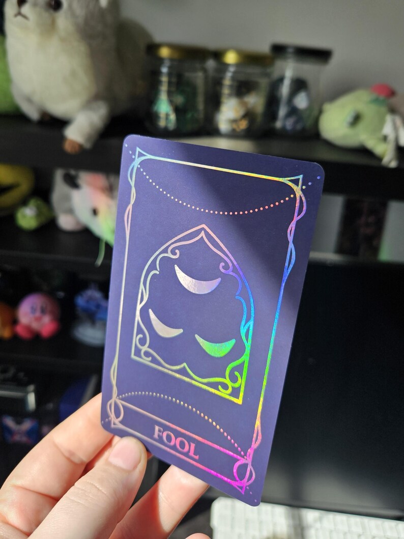 May include: A rectangular tarot card with a dark purple matte finish and holographic foil detailing. The card features an arched window design containing three crescent moon shapes, with the word FOOL printed in a rectangular frame at the bottom.