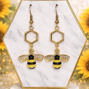 May include: A pair of gold-coloured bee earrings. Each earring features a black and yellow striped bee pendant with rhinestone wings, suspended from a gold hexagon. The earrings are displayed on a white and gold hexagonal stand.