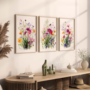 Op de afbeelding: Drie ingelijste aquarellen met kleurrijke bloemboeketten. De schilderijen hangen aan een witte muur boven een houten tafel met decoratieve items.