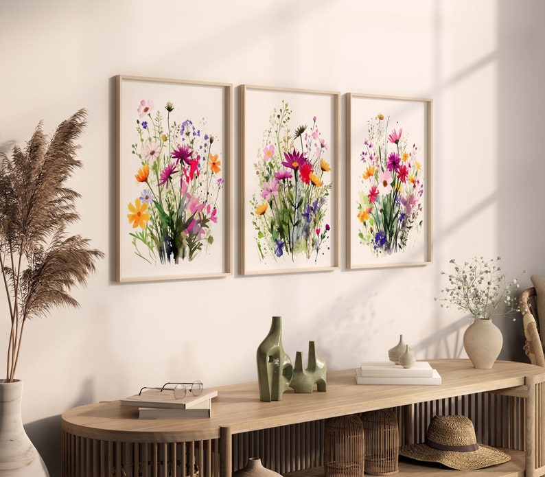 Op de afbeelding: Drie ingelijste aquarellen met kleurrijke bloemboeketten. De schilderijen hangen aan een witte muur boven een houten tafel met decoratieve items.