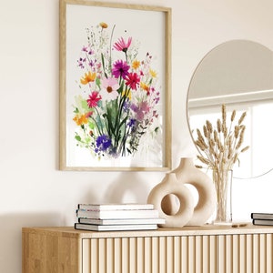 Op de afbeelding: Een ingelijst aquarel schilderij van een boeket kleurrijke bloemen in tinten roze, paars, geel en groen. Het schilderij hangt aan een witte muur boven een houten kast met een vaas en gedroogde bloemen.