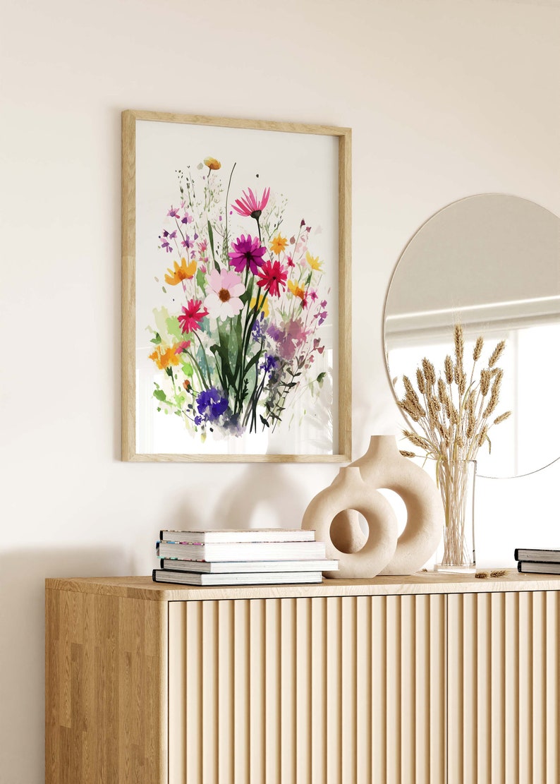 Op de afbeelding: Een ingelijst aquarel schilderij van een boeket kleurrijke bloemen in tinten roze, paars, geel en groen. Het schilderij hangt aan een witte muur boven een houten kast met een vaas en gedroogde bloemen.