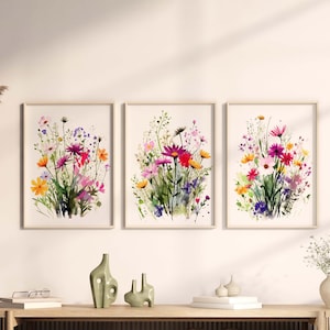 Op de afbeelding: Drie aquarellen van kleurrijke wilde bloemen in tinten roze, paars, geel en oranje. De schilderijen zijn ingelijst in eenvoudige houten lijsten en worden getoond op een witte muur.