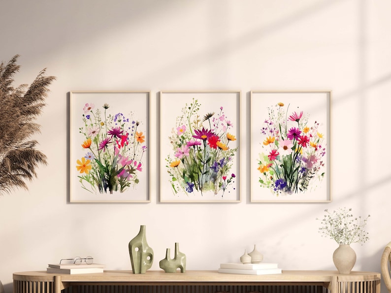 Op de afbeelding: Drie aquarellen van kleurrijke wilde bloemen in tinten roze, paars, geel en oranje. De schilderijen zijn ingelijst in eenvoudige houten lijsten en worden getoond op een witte muur.