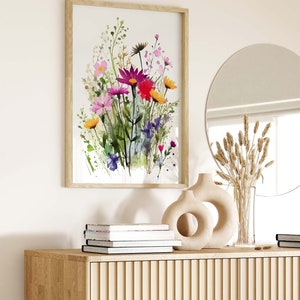 Op de afbeelding: Een ingelijst aquarel schilderij van een boeket kleurrijke wilde bloemen in tinten roze, paars, rood, geel en groen. Het schilderij hangt aan een witte muur boven een houten kast met een ronde spiegel en decoratieve vazen.