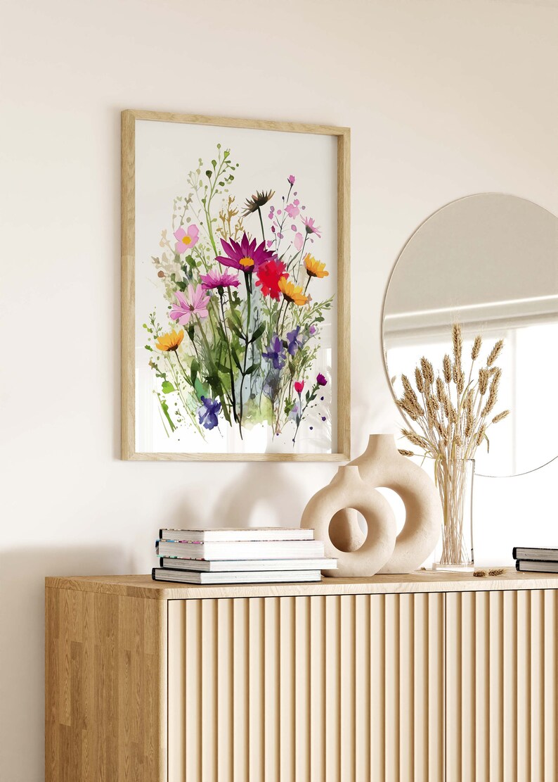 Op de afbeelding: Een ingelijst aquarel schilderij van een boeket kleurrijke wilde bloemen in tinten roze, paars, rood, geel en groen. Het schilderij hangt aan een witte muur boven een houten kast met een ronde spiegel en decoratieve vazen.