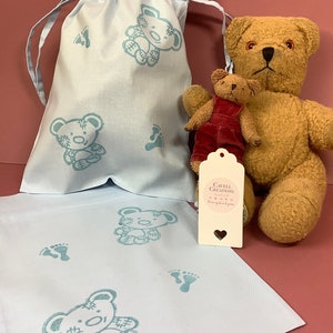 May include: A light blue fabric gift bag with a drawstring closure, featuring a repeated pattern of teddy bears and footprints. The bag is filled with two plush teddy bears, one brown and one red. A white gift tag with the text "Gavell Creations" is attached to the bag.