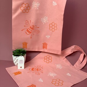 Puede incluir: Dos bolsas de lona rosa con un dise&ntilde;o de abeja y panal. Las bolsas tienen ilustraciones de abejas y panales blancas y naranjas. El texto en la bolsa m&aacute;s peque&ntilde;a dice "Craft Creations Handmade with Love".