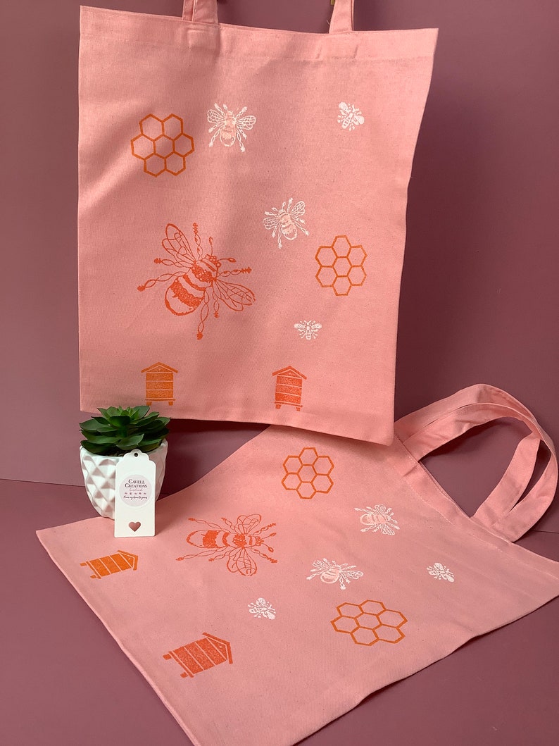 Puede incluir: Dos bolsas de lona rosa con un dise&ntilde;o de abeja y panal. Las bolsas tienen ilustraciones de abejas y panales blancas y naranjas. El texto en la bolsa m&aacute;s peque&ntilde;a dice "Craft Creations Handmade with Love".