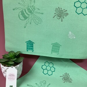 Puede incluir: Dos toallas de cocina verdes con un dise&ntilde;o de abeja y panal. Las toallas est&aacute;n dobladas y colocadas sobre una superficie rosa. Una peque&ntilde;a planta suculenta est&aacute; en una maceta blanca en primer plano. Una etiqueta blanca con el texto "Cavell Creations" est&aacute; unida a la maceta.