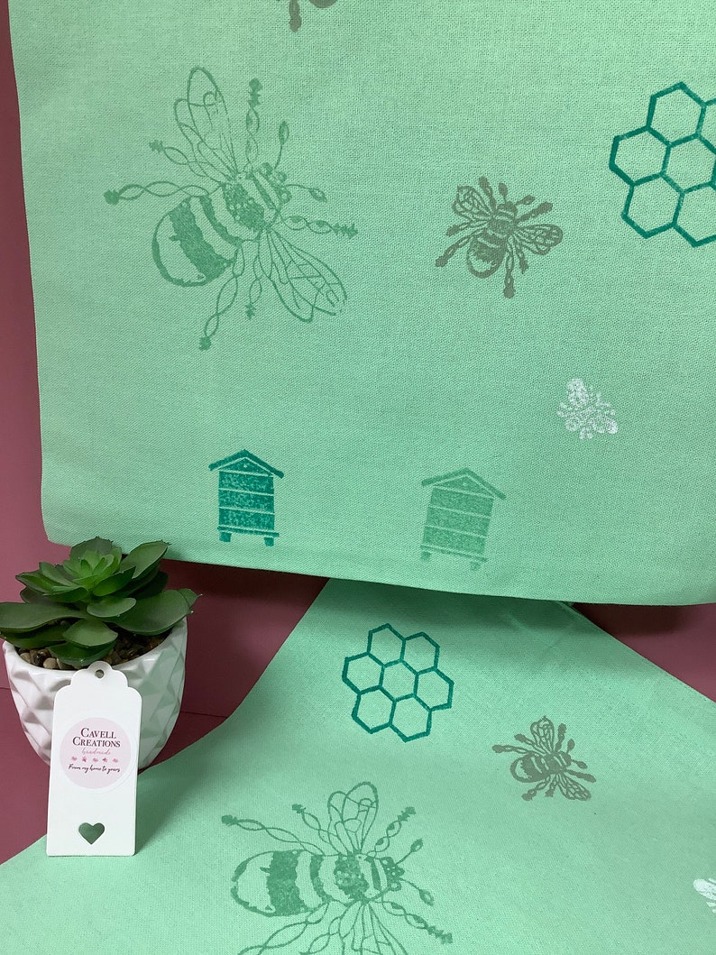 Puede incluir: Dos toallas de cocina verdes con un dise&ntilde;o de abeja y panal. Las toallas est&aacute;n dobladas y colocadas sobre una superficie rosa. Una peque&ntilde;a planta suculenta est&aacute; en una maceta blanca en primer plano. Una etiqueta blanca con el texto "Cavell Creations" est&aacute; unida a la maceta.