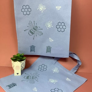 Puede incluir: Dos bolsas de tela azul claro con un dise&ntilde;o de abeja y panal. Las bolsas tienen dise&ntilde;os de abejas y panales blancos y grises. Las bolsas est&aacute;n hechas de una tela suave y natural.