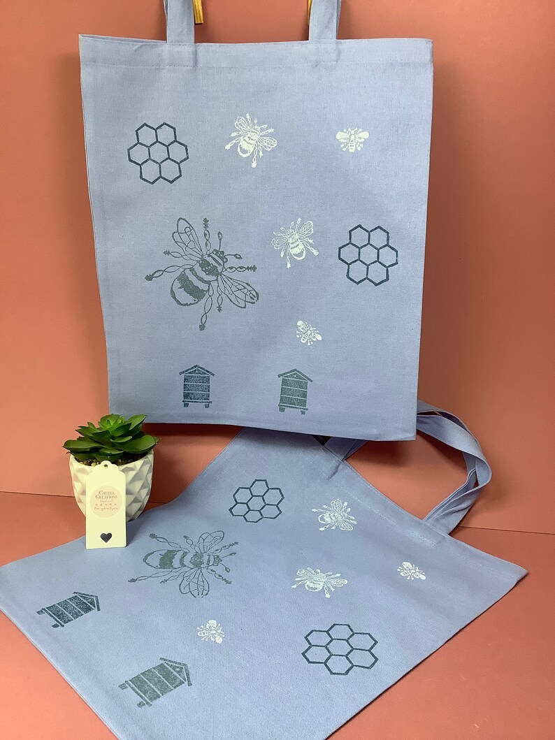 Puede incluir: Dos bolsas de tela azul claro con un dise&ntilde;o de abeja y panal. Las bolsas tienen dise&ntilde;os de abejas y panales blancos y grises. Las bolsas est&aacute;n hechas de una tela suave y natural.