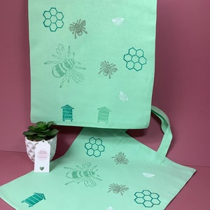 Puede incluir: Dos bolsas de lona verde con un dise&ntilde;o de abejas y panales. Las bolsas tienen asas blancas y est&aacute;n decoradas con un patr&oacute;n repetido de abejas, panales y colmenas.