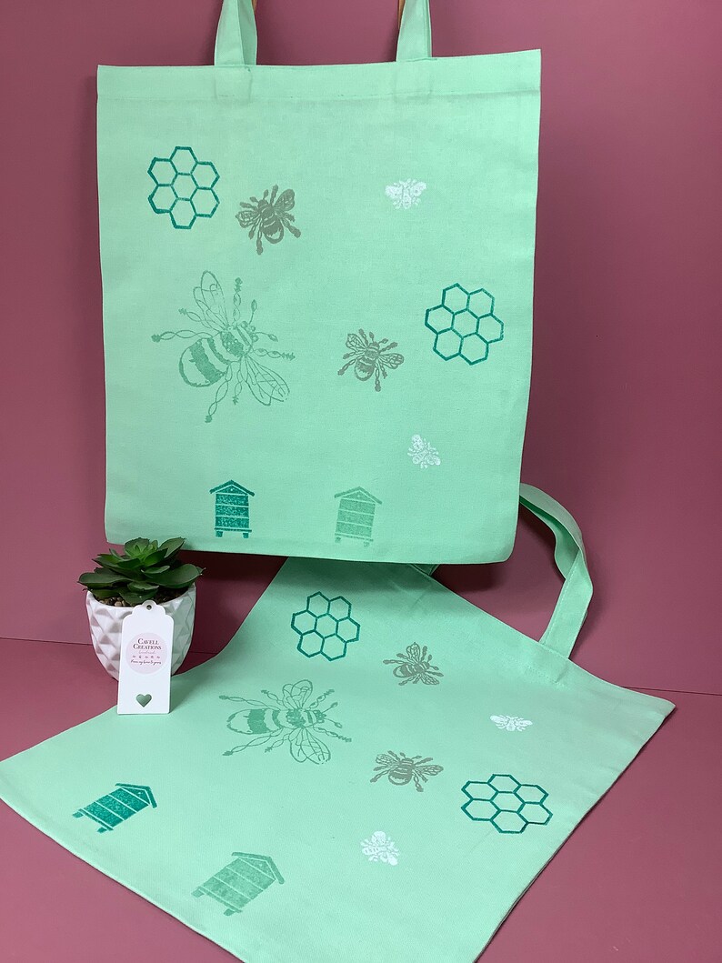 Puede incluir: Dos bolsas de lona verde con un dise&ntilde;o de abejas y panales. Las bolsas tienen asas blancas y est&aacute;n decoradas con un patr&oacute;n repetido de abejas, panales y colmenas.