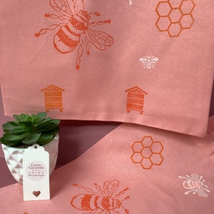 Puede incluir: Dos toallas de cocina rosas con un dise&ntilde;o de abeja y panal. Las toallas est&aacute;n dobladas y se muestran con una peque&ntilde;a planta suculenta en maceta y una etiqueta blanca que dice "Cavell Creations Handmade from my home to yours".