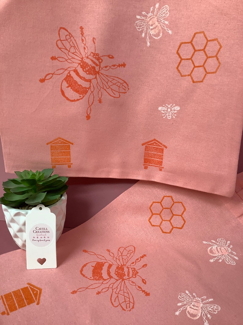 Puede incluir: Dos toallas de cocina rosas con un dise&ntilde;o de abeja y panal. Las toallas est&aacute;n dobladas y se muestran con una peque&ntilde;a planta suculenta en maceta y una etiqueta blanca que dice "Cavell Creations Handmade from my home to yours".
