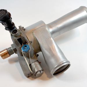 Może przedstawiać: Metalowy, srebrny element przemysłowy z cylindrycznym korpusem i różnymi mocowaniami. Posiada czarne złącze, miedziane złącze i niebieską nasadkę. Obiekt wykonany jest z metalu i umieszczony na białym tle.