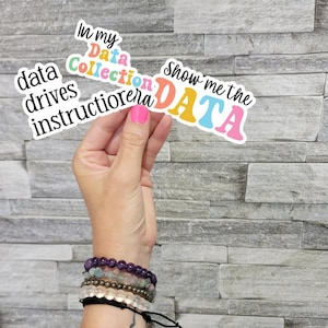 May include: A hand holding colourful stickers with phrases such as "Show me the DATA" and "In my Data Collection instructor era." The stickers have a white border and are in various colours. The person is wearing bracelets.