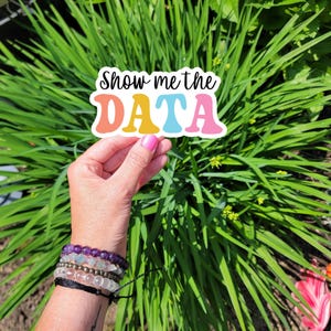 May include: A colourful sticker with the text "Show me the DATA" in a retro font. The sticker is held in a hand with several bracelets.