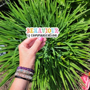 Può includere: Un adesivo con il testo "BEHAVIOUR is communication" in colori arcobaleno. L'adesivo è tenuto da una mano con un mucchio di braccialetti al polso.
