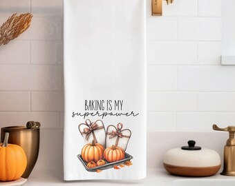 Può includere: Asciugamano da cucina bianco con la scritta "BAKING IS MY SUPERPOWER". L'asciugamano presenta un'illustrazione ad acquerello di zucche, guanti da forno e foglie d'autunno su una teglia. L'asciugamano è appeso a un'asta dorata.