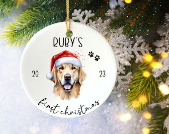 Puede incluir: Adorno navideño de cerámica blanca con un golden retriever con gorro de Papá Noel. El adorno tiene el texto "Ruby's first christmas" y el año "2023". Incluye huellas de patas y una cuerda dorada para colgar.