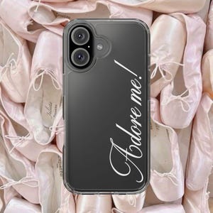 May include: Clear phone case with the text "Adore me!" in white script. The case is over a background of pink ballet shoes, creating a soft, feminine aesthetic. The phone case has a dual-lens camera.