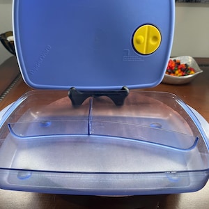 May include: A blue Tupperware container with a clear, divided interior. The lid is a darker blue with a yellow button and the Tupperware logo. The container is open, revealing the interior. The container is on a brown surface.