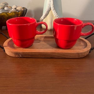 以下が含まれることがあります： 丸みを帯びたハンドルが付いた2つの鮮やかな赤いセラミックコーヒーマグが、黒い金属製のハンドルが付いた木製のサービングトレイに置かれています。マグカップは積み重ねられたデザインです。トレイは楕円形で、明るい色の木材で作られています。