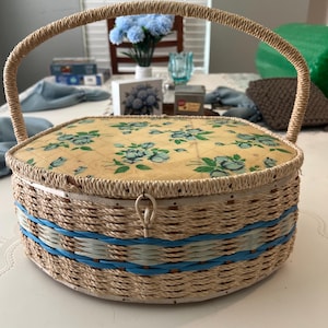 May include: A vintage sewing basket with a woven straw body and a hinged lid. The lid features a floral pattern in shades of blue and green on a yellow background. The basket has a woven handle and is accented with blue bands.