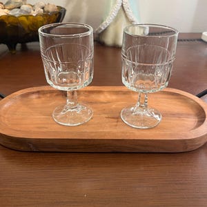 May include: Two clear, stemmed glasses with textured sides sit on a wooden serving tray. The glasses have a classic design, perfect for serving beverages. The tray is oval-shaped with a dark brown finish, adding a touch of elegance to the presentation.
