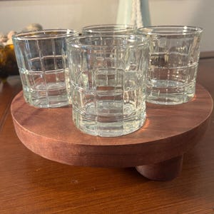 Puede incluir: Cuatro vasos transparentes con diseño cuadrado sobre una bandeja redonda de madera marrón oscuro. La bandeja tiene una base corta de pedestal. Los vasos están vacíos y parecen ser de vidrio.