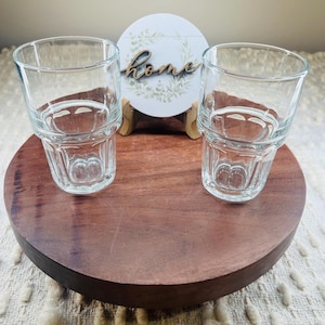 May include: Two clear, faceted drinking glasses sit on a round, dark wood serving tray. A small, round sign with the word "home" in cursive is behind the glasses. The sign has a floral design and is propped up on a small wooden stand.