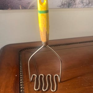 Puede incluir: Un antiguo pasapurés con mango de madera amarillo y una franja verde. El cabezal metálico del pasapurés tiene un diseño ondulado. El mango tiene un agujero para colgarlo. El utensilio de cocina está diseñado para machacar patatas y otros alimentos blandos.
