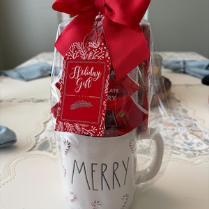 Può includere: Una tazza in ceramica bianca con la parola MERRY stampata in nero e piccoli motivi a bastoncino di zucchero rosso, riempita di cioccolato e avvolta in plastica trasparente con un grande fiocco rosso e un'etichetta rossa con scritto Holiday Gift.