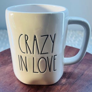 Peut inclure: Une tasse en céramique blanche avec les mots "CRAZY IN LOVE" en lettres noires. La tasse a une anse incurvée et repose sur une surface en bois. La tasse est de couleur neutre et a un design simple et moderne.