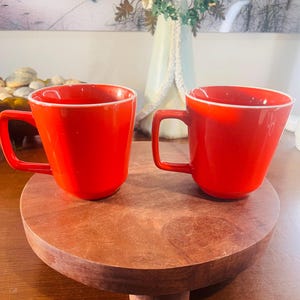 Puede incluir: Dos tazas rojas brillantes con bordes blancos sobre un pedestal de madera redondo. Las tazas tienen asas cuadradas y un acabado brillante. El pedestal es de color marrón cálido.