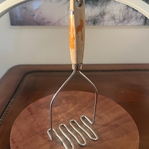 May include: A potato masher with a wooden handle and a stainless steel mashing head. The handle has orange accents. The masher is resting on a round, wooden cutting board. The brand name "NEEWER" is visible on the top.
