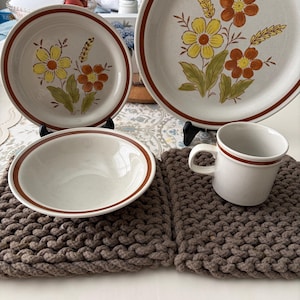 Può includere: Un set di stoviglie in ceramica con finitura bianco sporco maculato e bordi marroni. Il set comprende un piatto piano grande, un piatto più piccolo, una ciotola poco profonda e una tazza, tutti decorati con motivi floreali gialli e arancioni.