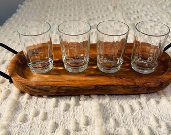 May include: Four clear glass shot glasses with a ribbed design are arranged on a wooden tray with black metal handles.