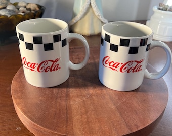 Op de afbeelding: Twee witte Coca-Cola keramische mokken met zwart-witte geblokte banden en het Coca-Cola logo in rood. De mokken staan op een ronde, houten serveerschaal.