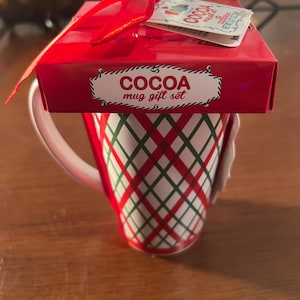 May include: A white ceramic mug with a black handle and the text "But First, Coffee" in a bold, black font. The mug is set against a blurred background, suggesting a cozy setting.