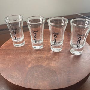 May include: Four clear shot glasses with the text "Sauza 901" printed in black. The shot glasses are arranged on a round, dark brown wooden tray. The shot glasses are empty and ready to be filled.