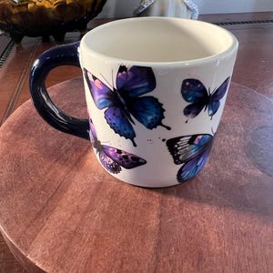 May include: White ceramic mug with a dark blue handle. The mug is decorated with purple and blue butterfly illustrations. The mug is sitting on a round, wooden surface.