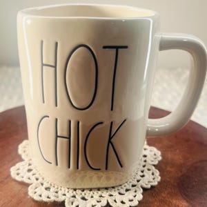 Peut inclure: Mug en céramique crème avec les mots "HOT CHICK" en lettres noires. La tasse a une poignée incurvée et repose sur un dessous de verre en dentelle crème. La tasse est sur une surface en bois brun foncé.