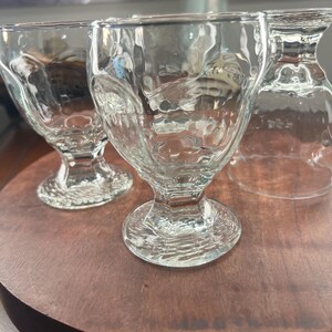 Op de afbeelding: Drie heldere glazen dessertbekers met getextureerde zijkanten en een voetstuk staan op een ronde houten schaal. De bekers hebben een unieke, gehamerde glazen uitstraling, die licht reflecteert en de presentatie verbetert.