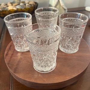 Puede incluir: Cuatro vasos de chupito transparentes con diseños colocados sobre una bandeja redonda de madera. Los vasos tienen un diseño detallado y texturizado. La bandeja es de color marrón cálido y los vasos están vacíos.